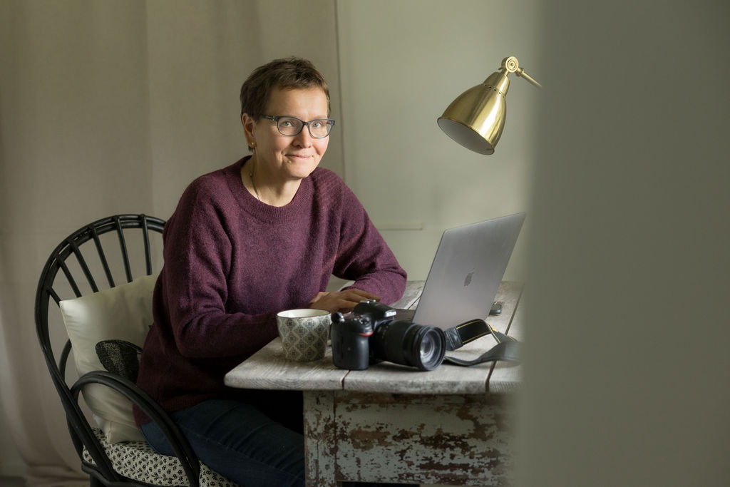 Nainen istuu työpöydän ääressä, edessä on läppäri, kamera ja kahvimuki.